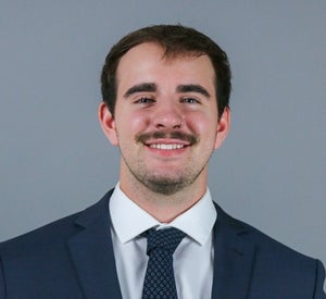 Man with short brown hair and a mustache wearing a navy blue suit jacket and a white collared shirt with a navy blue and black tie.