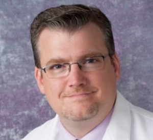 A man with brown hair and glasses wearing a white lab coat over a light pink collared shirt.