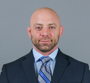 Bald man wearing a navy blue suit jacket over a light blue collared shirt and a blue and gray striped tie.