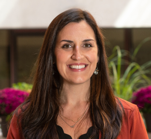 Woman with long brown hair wearing an orange blouse over a black shirt.