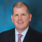 Man with very short light brown hair wearing a black suit jacket over a light blue collared shirt and a red tie.