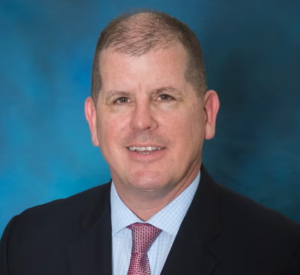 Man with very short light brown hair wearing a black suit jacket over a light blue collared shirt and a red tie.