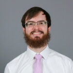 Man with red/brown hair and a beard wearing a white collared shirt and a light pink tie.