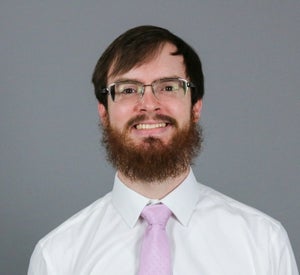 Man with red/brown hair and a beard wearing a white collared shirt and a light pink tie.