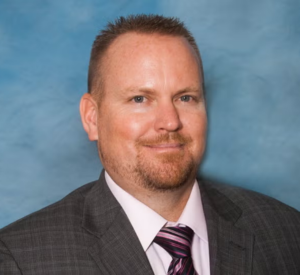 Man with short auburn hair wearing a brown suit jacket over a white collared shirt and a pink and black striped tie.