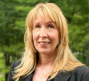 Woman with blonde hair wearing a dark gray blazer over a black and white polka dot blouse.