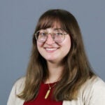 Woman with medium length, brown hair with bangs wearing wire glasses and a red top with beige cardigan.