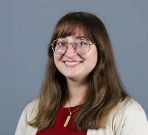 Woman with medium length, brown hair with bangs wearing wire glasses and a red top with beige cardigan.