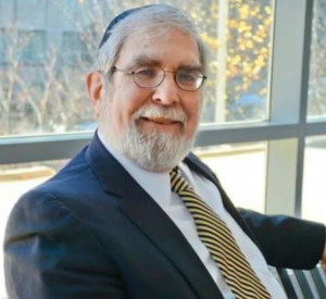 Man with white hair and a beard wearing a black suit jacket over a white collared shirt and a black and yellow striped tie.