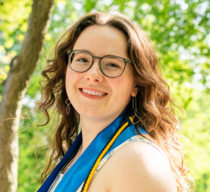 Woman with brown curly hair wearing glasses and a white top with a blue ribbon and a yellow graduation cord around her neck.