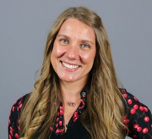 Woman with long, light brown hair pulled back wearing a black blouse with red cherries printed on it and a silver necklace.