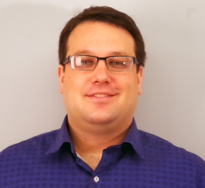 Man with short brown hair wearing a dark blue collared shirt with black squares and black rimmed glasses.