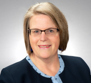 Woman with short, dark blonde hair wearing a black blazer over a light blue shirt.