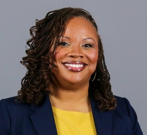 Woman with dark brown curly hair wearing a navy blue blazer over a yellow top.