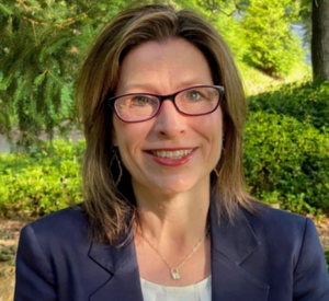 Woman with short brown and blonde hair wearing a navy blue blazer over a white blouse with a silver necklace and dark glasses.