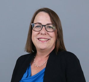 Woman with short brown hair wearing black glasses, a silver necklace and a black sweater over a dark blue top.