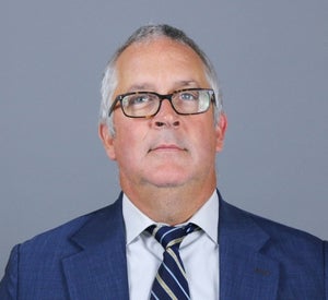 Man with short gray hair wearing brown glasses and a blue suit jacket over a white collared shirt and a dark blue and gold striped tie.