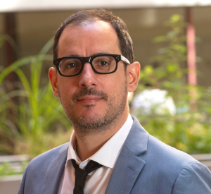 Man with short brown hair wearing black framed glasses and a light gray suit jacket over a white collared shirt and a black tie.