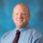 Man with very short hair wearing a blue and white checker patterned collared shirt with a dark red tie.