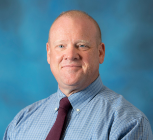 Man with very short hair wearing a blue and white checker patterned collared shirt with a dark red tie.