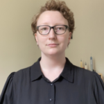 A woman with very short curly hair wearing a black collared blouse and black glasses.