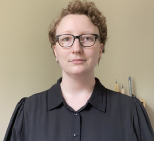 A woman with very short curly hair wearing a black collared blouse and black glasses.