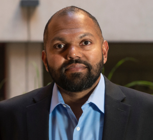 Man with very short black hair and a beard wearing a black suit jacket over a light blue collared shirt.