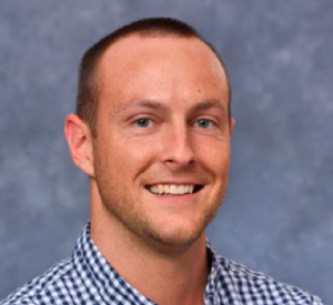 Man with very short brown hair wearing a black and white checkered, collared shirt.