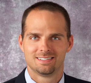 Man with short dark hair wearing a black suit jacket over a light gray collared shirt.