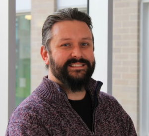 Man with dark hair and a beard wearing a purple sweater over a black shirt.