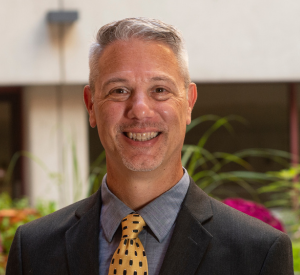 Man with short gray hair wearing a black suit jacket over a dark gray collared shirt and a gold tie with dark blue squares.