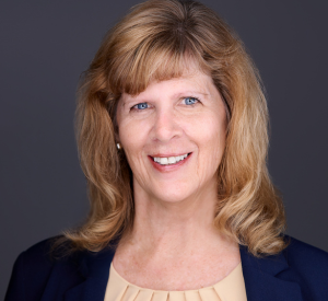 Woman with dark blonde hair wearing a black blazer over a tan blouse.