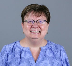 Woman with short brown hair wearing black rimmed glasses and a purple blouse with floral print.