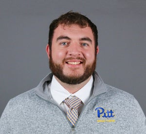 Man with short brown hair and a beard wearing a gray Pitt pullover over a white collared shirt and a blue and yellow striped tie.