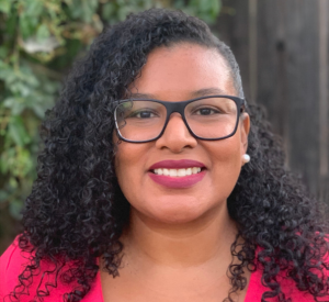 Woman with dark curly hair and dark glasses wearing a bright pink shirt.