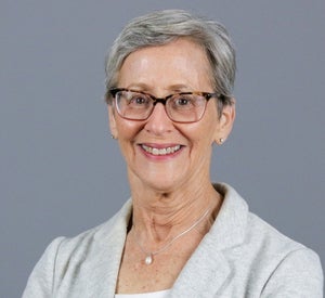 Woman with short gray hair wearing a cream blazer over a white top, black and light pink glasses and a silver pearl necklace.