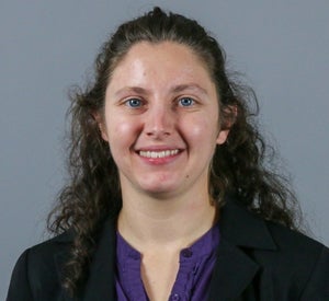 Woman with long brown curly hair pulled back wearing a black blazer over a dark purple top.