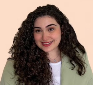 A woman with long, brown curly hair wearing a light green blazer over a white shirt.