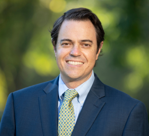 Man with short black hair wearing a navy blue suit jacket over a light blue collared shirt and a yellow and blue patterned tie.