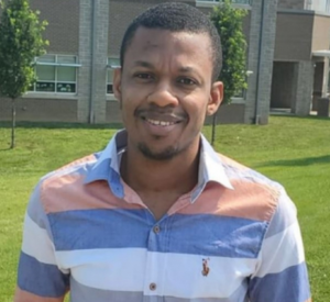 Man with short dark hair waring a collared shirt with white, gray, blue and peach stripes.