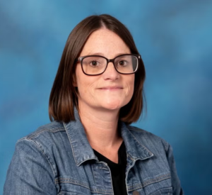 A woman with, short dark auburn hair wearing a denim jacket over a black shirt.