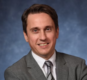 Man with brown hair wearing a dark gray blazer over a white collared shirt with a silver tie.