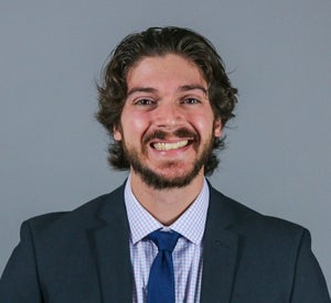 Man with brown curly hair and a beard wearing a navy blue suit over a white and blue checkered shirt and a dark blue tie.