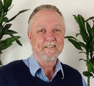 Man with very short gray hair wearing a blue and white striped collared shirt under a black sweater vest.