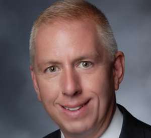 Man with very short white hair wearing a black suit jacket over a white collared shirt.