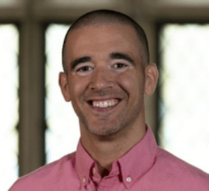 Man with very short dark hair wearing a pink collared shirt.