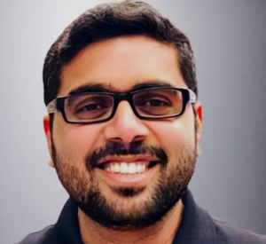 Man with dark hair and a beard wearing glasses and a dark gray collared shirt.