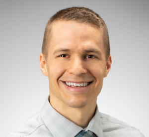 Man with short blonde hair wearing a light gray collared shirt and a blue tie.