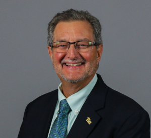 Man with dark brown and gray hair wearing a black suit jacket over a light green collared shirt with a teal patterned tie and a Pitt lapel pin.