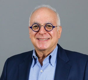 Man with short white hair and black circular glasses wearing a navy blue suit jacket over a light blue collared shirt.